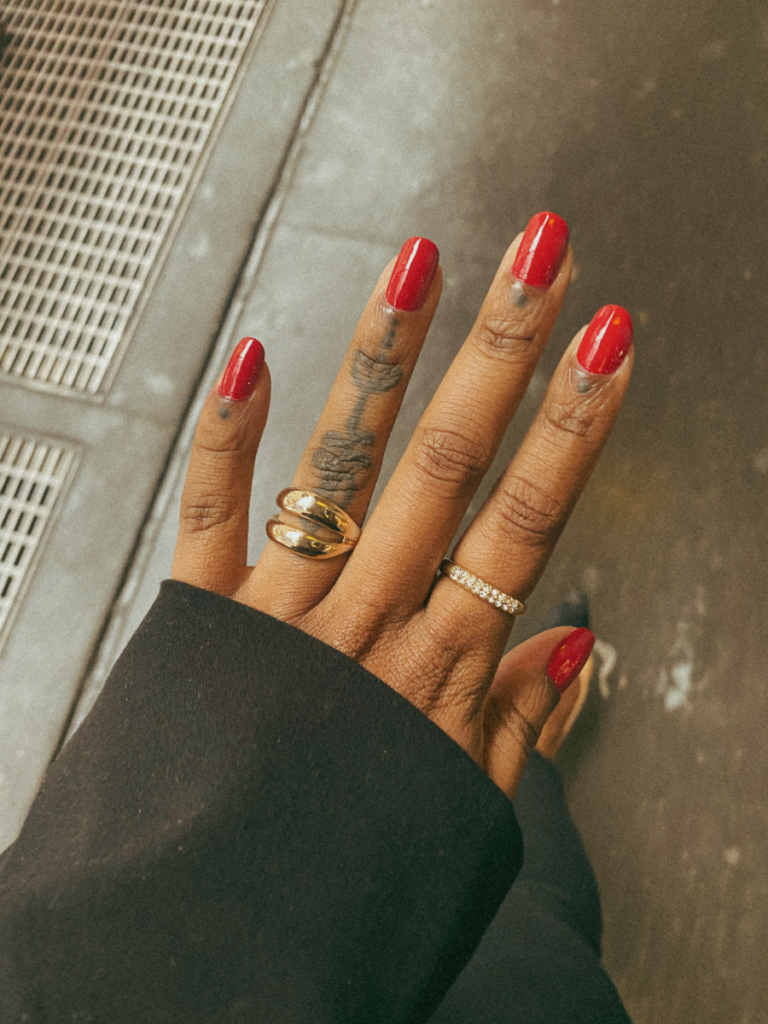 A black woman's hand with a freshly painted red manicure using Chanel Le Vernis, and wearing two gold rings with a black blazer sleeve in view.