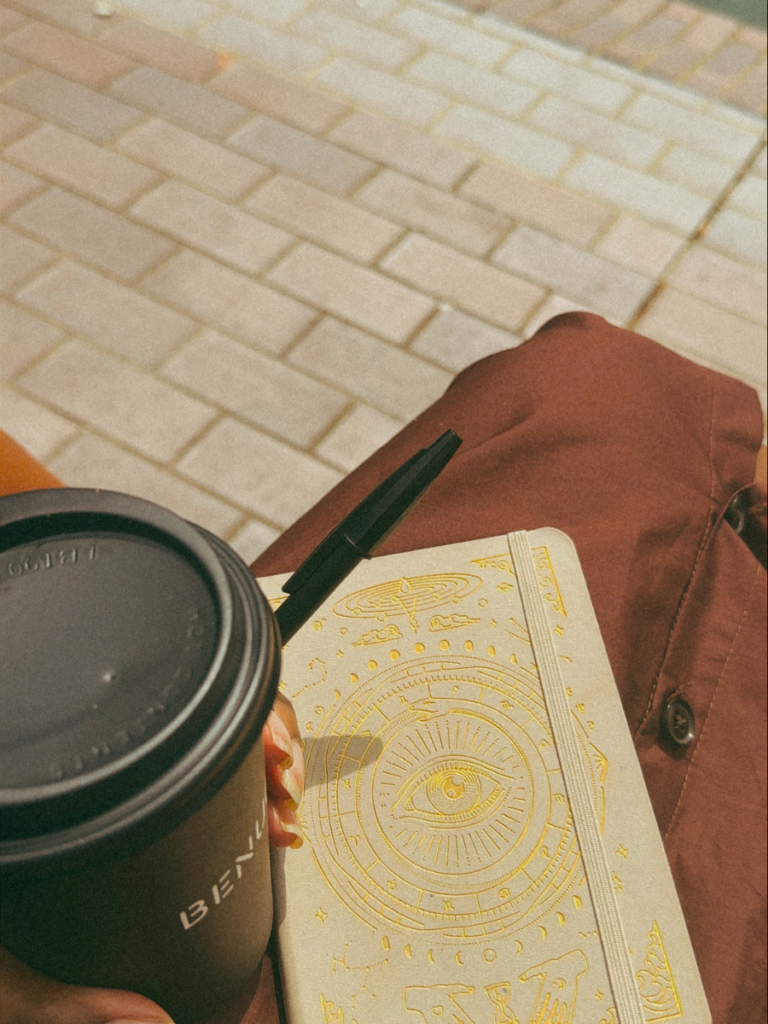 A Black woman's hand holding a takeaway coffee from Benugo with a pen and journal from Magic of I, wearing a brown shirt dress while sitting on a bench outside of Barbican, London.