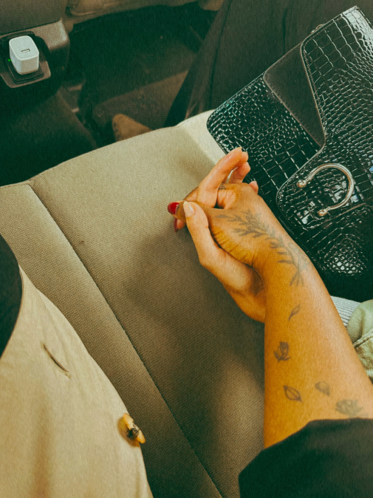 Two friends holding hands, one black woman and white woman, one with tattoos, in the back of a car, with a black mock crocodilehandbag